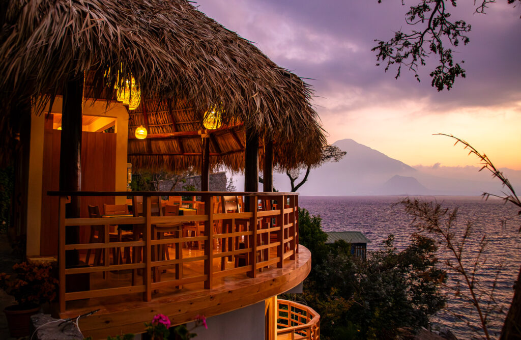 B'alam Ya Cafe view: Thatched roof palapa overlooking Lake Atitlán at sunset with a volcano backdrop.